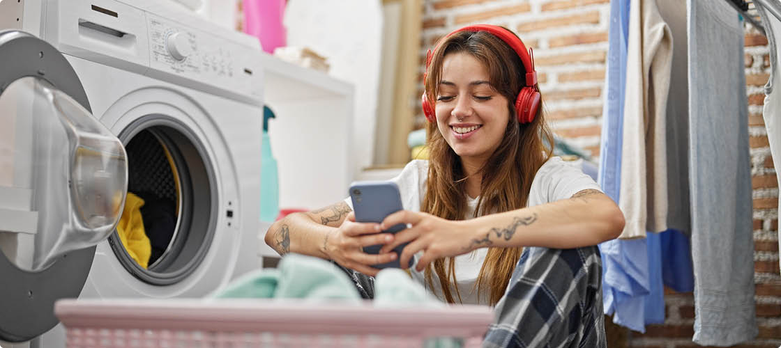 Young beautiful hispanic woman using smartphone and headphones washing clothes at laundry room